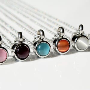 Può includere: Una collezione di collane d'argento, ognuna con un ciondolo rotondo con una pietra preziosa di diversi colori. Le pietre preziose includono rosa, viola, turchese, arancione, bianco e verde. I ciondoli sono incastonati in una lunetta d'argento.