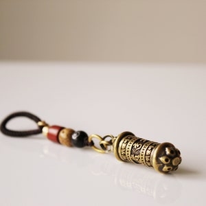 May include: A brown leather cord with wooden beads and a brass charm with engraved text. The charm is shaped like a small cylinder with a decorative top.