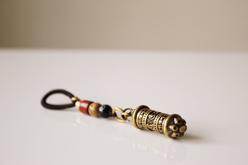 May include: A brown leather cord with wooden beads and a brass charm with engraved text. The charm is shaped like a small cylinder with a decorative top.