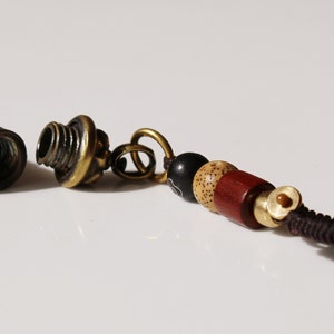 May include: A small, brass, cylindrical container with intricate engravings, open and revealing an empty interior. It is attached to a brown cord with a small, brass bead and a series of wooden beads in black, beige, and red.