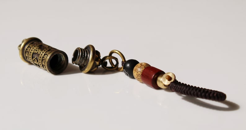 May include: A small, brass, cylindrical container with intricate engravings, open and revealing an empty interior. It is attached to a brown cord with a small, brass bead and a series of wooden beads in black, beige, and red.