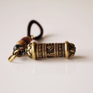 May include: A gold-colored metal prayer wheel with intricate designs and lettering. The wheel is attached to a brown cord with beads.