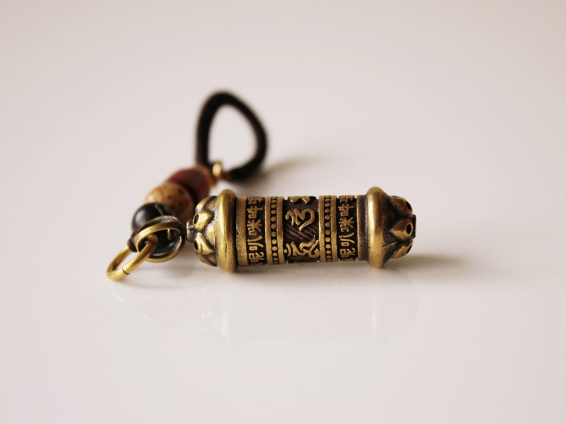 May include: A gold-colored metal prayer wheel with intricate designs and lettering. The wheel is attached to a brown cord with beads.