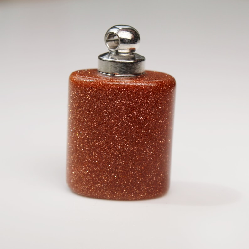May include: A small, oval-shaped, brown, glittery bottle pendant with a silver cap and a loop for attaching to a chain.