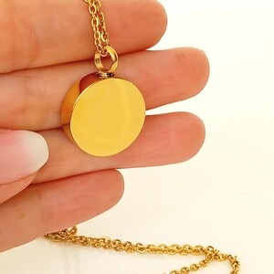 May include: A gold-colored pendant necklace featuring a round, polished charm. The charm is attached to a delicate chain. The necklace is held in a hand against a white background. The pendant has a smooth, reflective surface.