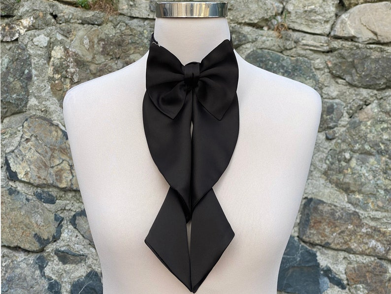 May include: A black satin bow tie with a long, flowing design. The bow tie is tied around a white mannequin's neck.