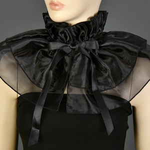 May include: A black ruffled collar with a high neck and a large bow. The collar is made of a shiny, sheer fabric and is worn over a black top. The design is reminiscent of vintage fashion.