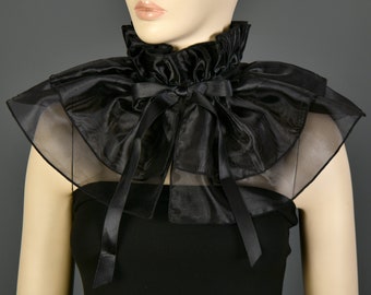 May include: A black ruffled collar with a high neck and a large bow. The collar is made of a shiny, sheer fabric and is worn over a black top. The design is reminiscent of vintage fashion.