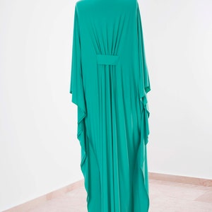 May include: A long, flowing teal-colored dress with wide sleeves is displayed on a black mannequin. The dress features a gathered detail at the waist and a simple neckline. The fabric drapes elegantly, showcasing its fluid design.