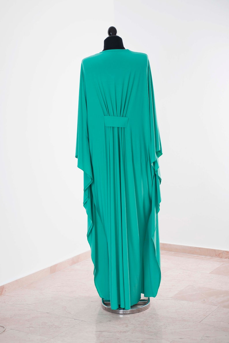 May include: A long, flowing teal-colored dress with wide sleeves is displayed on a black mannequin. The dress features a gathered detail at the waist and a simple neckline. The fabric drapes elegantly, showcasing its fluid design.