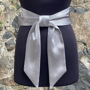 Puede incluir: Una cinta de satén plateada atada en un lazo alrededor de una cintura. La cinta es larga y fluida, con los extremos de la cinta apuntando hacia abajo.