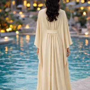 May include: A long, flowing, cream-colored dress with wide sleeves and a belted waist. The dress is worn by a person standing near a pool of water. The background features a building with soft lighting.