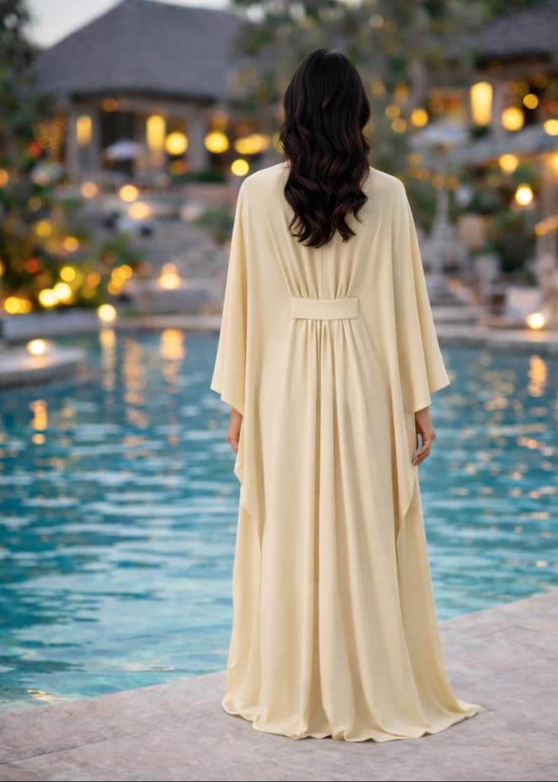 May include: A long, flowing, cream-colored dress with wide sleeves and a belted waist. The dress is worn by a person standing near a pool of water. The background features a building with soft lighting.