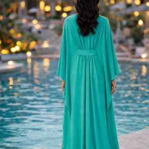 May include: A long, flowing turquoise dress with wide sleeves and a cinched waist. The dress is made of a lightweight fabric and is worn by a person standing near a pool. The background features a blurred view of a building and lights.