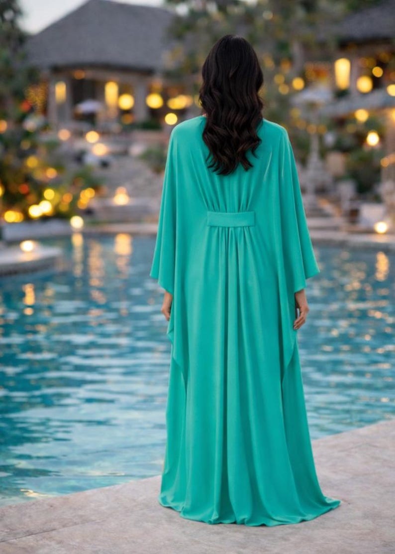 May include: A long, flowing turquoise dress with wide sleeves and a cinched waist. The dress is made of a lightweight fabric and is worn by a person standing near a pool. The background features a blurred view of a building and lights.