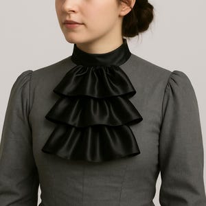 May include: A black satin ruffled collar with three tiers, worn over a gray dress. The collar has a high neckline and is a striking contrast against the dress. The ruffles add a touch of elegance.