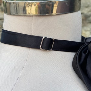 May include: A black satin bow tie with a silver metal buckle. The bow tie is on a white mannequin.