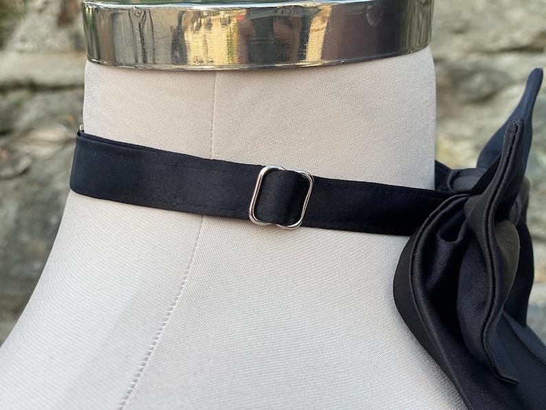 May include: A black satin bow tie with a silver metal buckle. The bow tie is on a white mannequin.