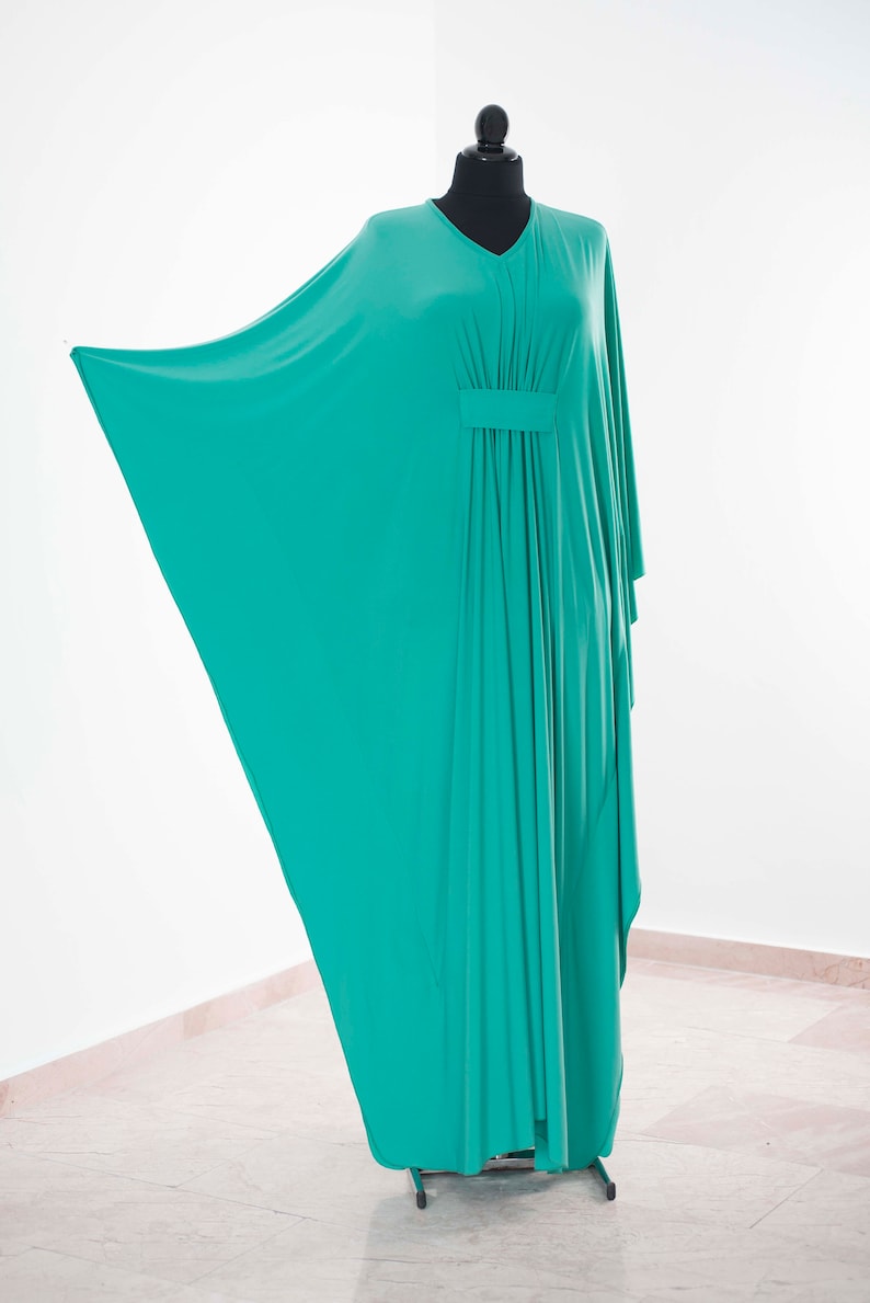 May include: A teal-colored, flowing kaftan dress displayed on a black mannequin. The dress features a V-neckline and a gathered detail at the waist. The fabric drapes elegantly, showcasing its loose, comfortable design. The dress is made of a soft, lightweight material.