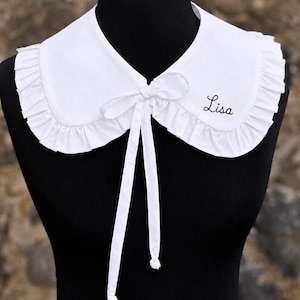 May include: A white detachable collar featuring a rounded Peter Pan style, ruffled edges, and a front tie closure. The fabric is plain white, and the name Lisa is embroidered in black cursive script on the right side of the collar piece.
