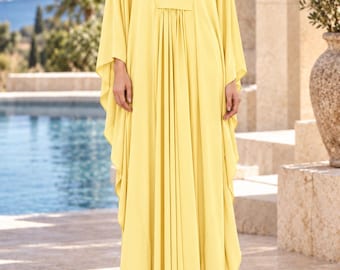 May include: A floor-length, pale yellow kaftan dress featuring a V-neckline, wide flowing sleeves, and a central rectangular panel at the waist with vertical pleats that extend down the length of the skirt, creating a draped, loose silhouette.