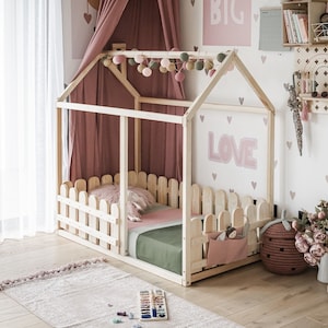 Peut inclure: Un lit en bois en forme de maison de jeu avec une canopée rose et une clôture en bois blanc autour du matelas. Le lit est décoré de cœurs roses et du mot "LOVE" en lettres roses.