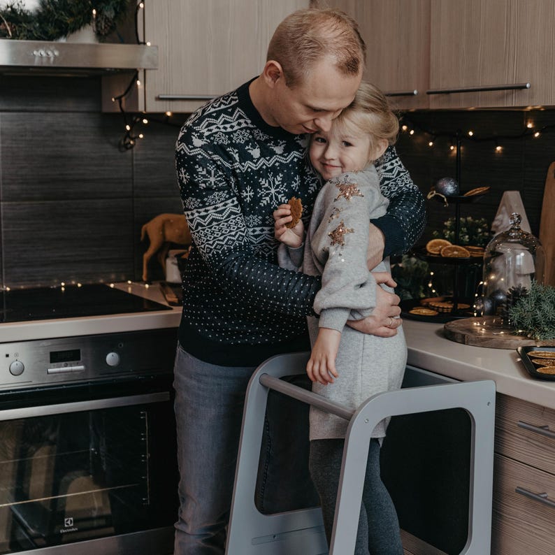 Father embracing his daughter standing on a grey montessori step stool during Christmas morning breakfast prep.