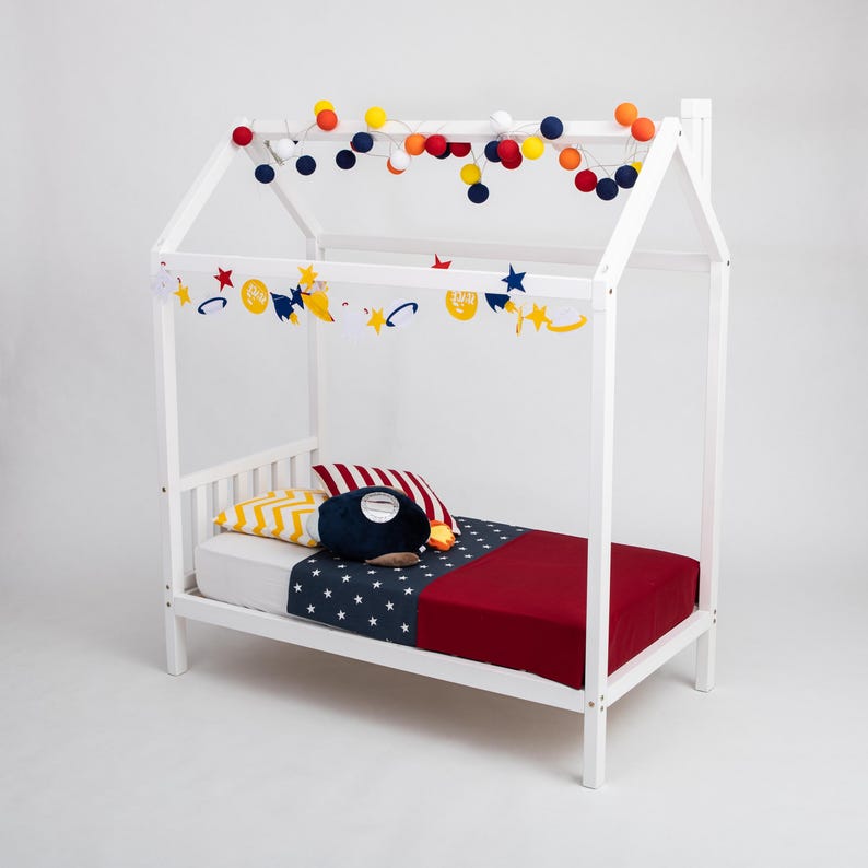 White toddler house bed on legs dressed with colorful ball string lights, red blanket, and patterned accent pillows.