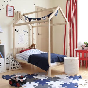 Side perspective of the wooden house bed on legs decorated with navy blanket, star banner, and wooden floor puzzle tiles.