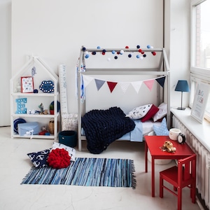 Toddler wooden house bed on legs positioned in a bright modern playroom setting with bookshelves, small red table, and rug.