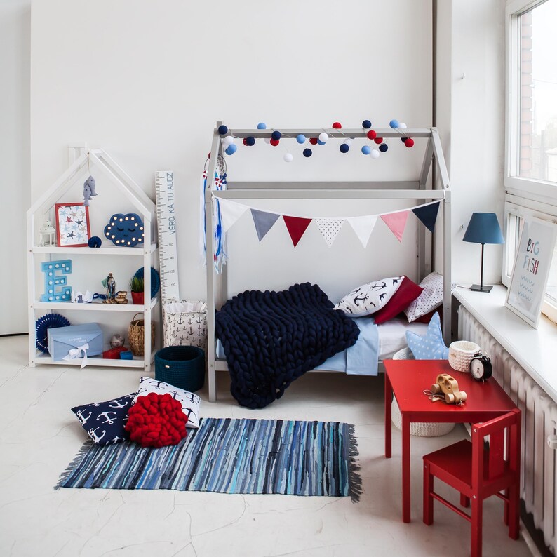 Toddler wooden house bed on legs positioned in a bright modern playroom setting with bookshelves, small red table, and rug.