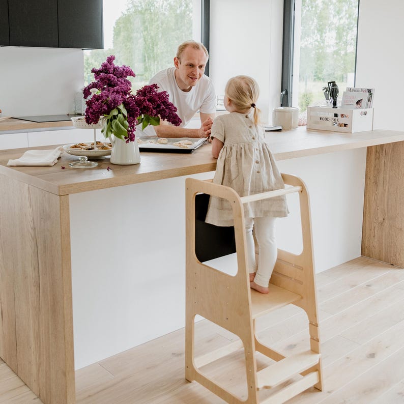 Little girl standing on a lacquered birch plywood kitchen step stool next to fresh flowers, reaching the counter safely.