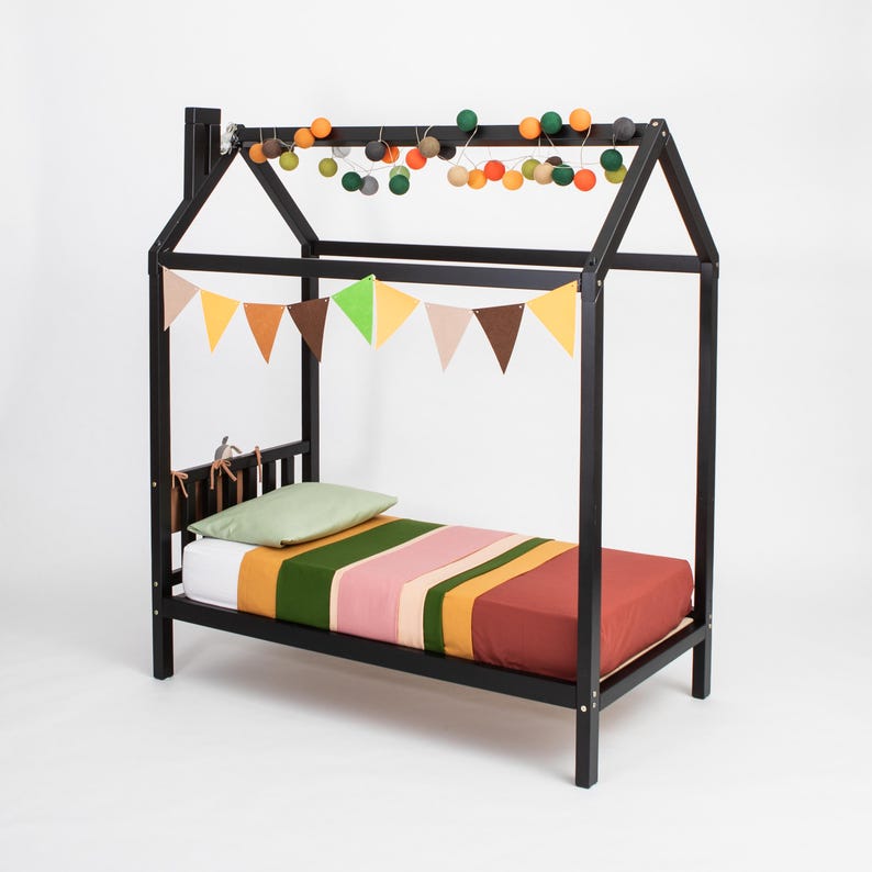 Black wooden house bed frame on raised legs featuring built-in mattress slats, decorated with hanging party buntings.