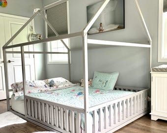 May include: A light gray wooden house-shaped bed frame with a peaked roof structure. The bed features vertical slat railings on the sides and is dressed with a light blue quilted blanket, a matching pillow, and a white patterned sheet set.