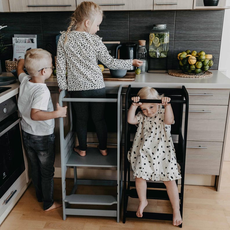 Three young children using grey and black adjustable step stools at the kitchen counter, ideal for growing families.