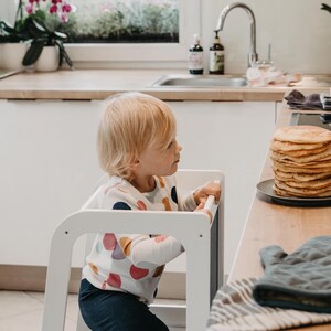 Young toddler standing on a white kitchen booster stool to reach snacks, solving daily height reach struggles.