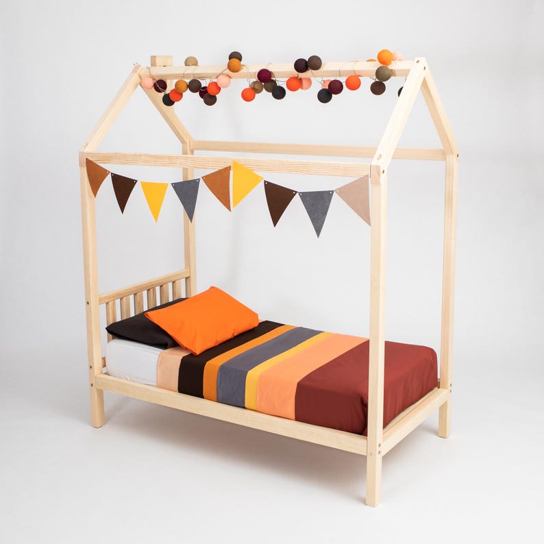 Solid wood house bed on legs with pom-pom roof bunting and warm autumn-striped bedsheets against a neutral studio backdrop.