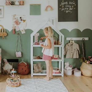 Puede incluir: Una estanter&iacute;a de madera blanca con forma de casita de mu&ntilde;ecas con tres estantes. Los estantes est&aacute;n llenos de juguetes, incluyendo una letra I rosa, un arco&iacute;ris, un collar de cuentas de madera y un tel&eacute;fono de juguete rosa. Una ni&ntilde;a est&aacute; de pie frente a la estanter&iacute;a, sosteniendo un juguete rosa. La estanter&iacute;a est&aacute; en una habitaci&oacute;n con paredes verdes y una alfombra blanca.