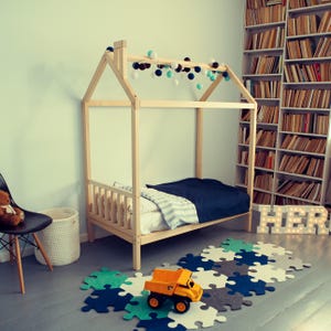 Raised natural pine house bed frame in a play area with soft green walls, alphabet blocks, and geometric floor mats.
