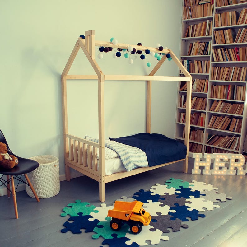 Raised natural pine house bed frame in a play area with soft green walls, alphabet blocks, and geometric floor mats.