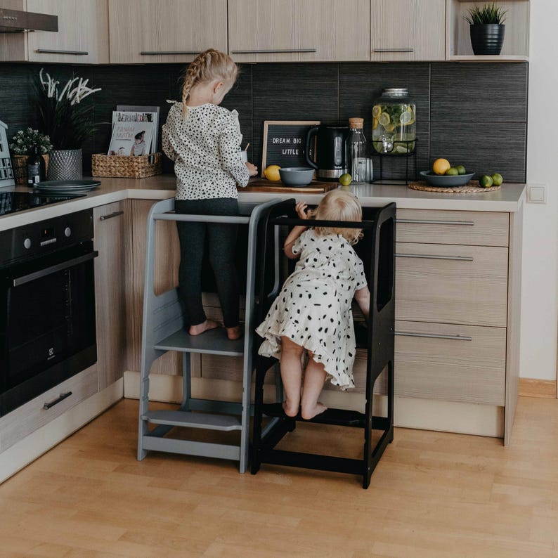 Two young girls standing on grey and black kitchen step stools, eliminating chair climbing during baking activities.