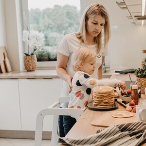 Mother helping her toddler make pancakes on a white step stool with a drawing board, promoting Montessori self-reliance.