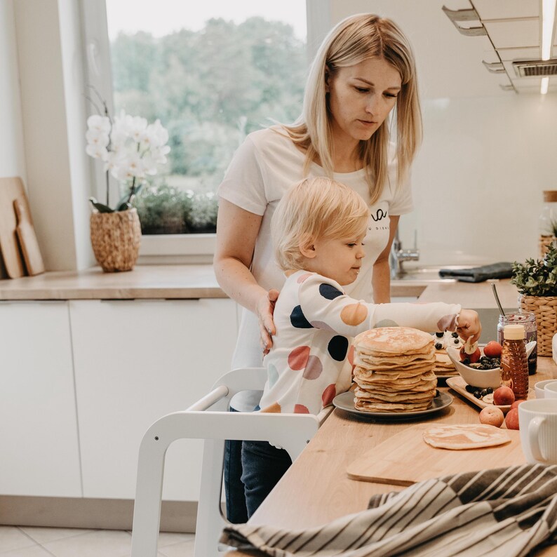 Mother helping her toddler make pancakes on a white step stool with a drawing board, promoting Montessori self-reliance.