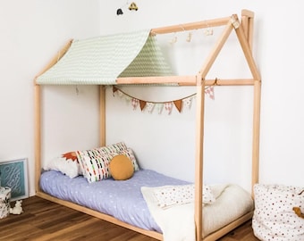 May include: A light-colored wooden house-shaped bed frame with a pale green patterned fabric roof. The bed features a light purple patterned sheet, a round mustard yellow pillow, a white blanket, and a decorative pennant banner hanging inside.
