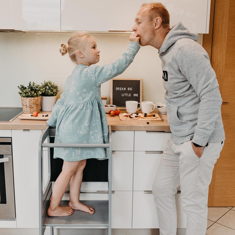 Little girl in a green dress sharing food with her father while standing on a grey child counter booster stool.