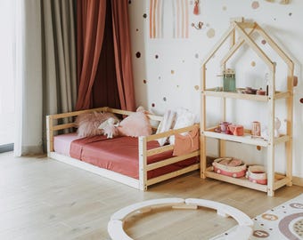 May include: A low wooden floor bed with a terracotta-colored sheet and pillows, positioned next to a three-tiered wooden house-shaped shelving unit. A circular wooden balance beam toy lies on the floor in front of the bed and shelf.