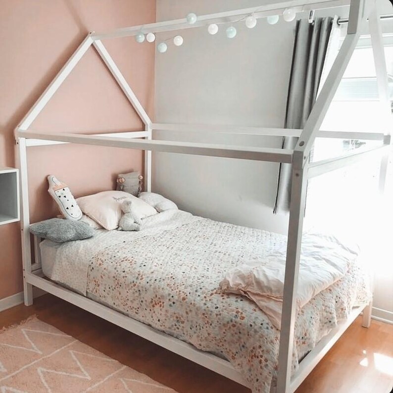 White wooden Montessori house bed on low legs placed against a pastel pink wall in a toddler girl bedroom with warm fairy lights.