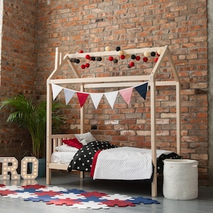 Natural wooden house bed on legs with slatted base, styled in an industrial-style child bedroom with loft brick wall and garland decor.
