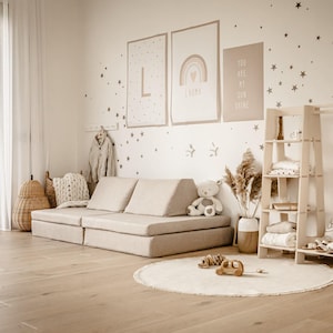Può includere: Una stanza dei giochi per bambini dai toni neutri con un divano modulare, scaffalature in legno e decorazioni murali. Un orsacchiotto bianco è seduto sul divano. La stanza presenta un tappeto rotondo, giocattoli di legno e decalcomanie a forma di stella.