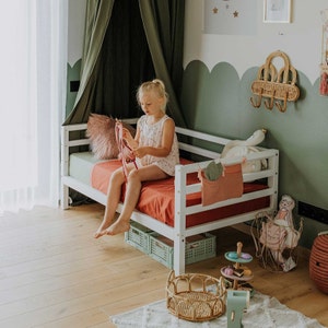 May include: A white wooden bed with a red mattress and a green and pink pillow. A young girl sits on the bed. The room features a green canopy, a woven wall hanging, and toys, including a swan plush and a small wooden toy set.